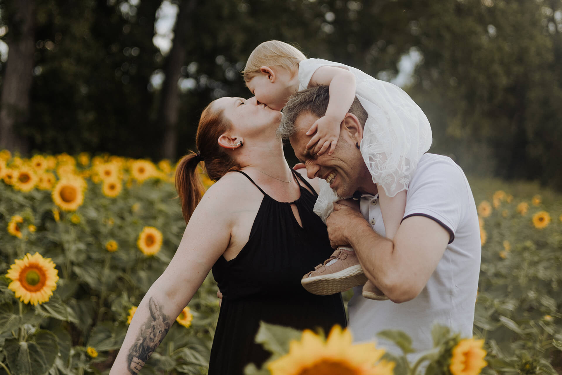 Kind sitzt auf Schultern vom Vater und gibt der Mutter einen Kuss